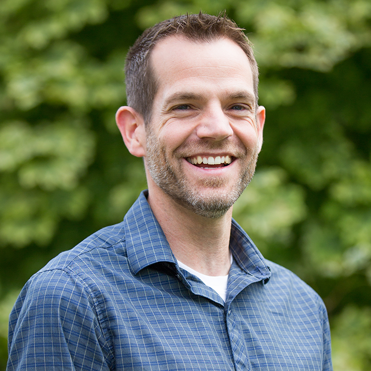 Portrait of Dr. Matthew Chatfield. He is smiling at the camera and wears a blue plaid shirt with a collar. He is standing outside in front of greenery. 