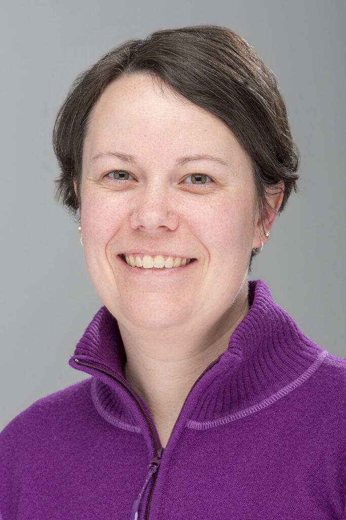 A portrait of Associate Dean Leahy. She wears a purple have zip sweater and has short dark hair. She is smiling at the camera. 