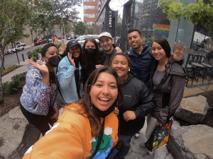 Eight people pose for a group photo outside in an urban environment. The person in the foreground is taking a selfie.