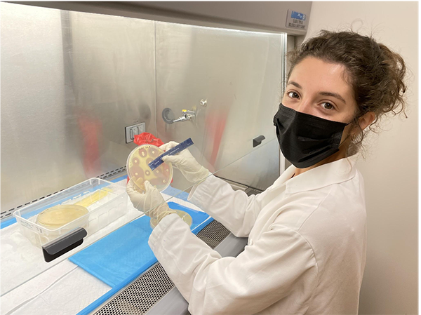 Roadcap smiles at the camera behind a mask. She is holding a petri dish and standing in front of a vent hood system in a laboratory.