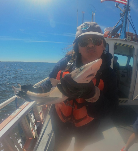 Pérez Santos pauses with a spiny dogfish shark to pose for a photo. She is standing on a boat wearing a hat, sunglasses, and orange PFD.