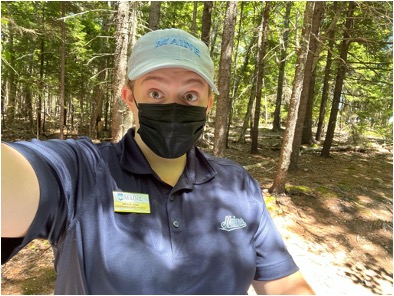The authors takes a selfie. She is wearing a green ballcap, a navy blue polo with a nametag and a black mask. Trees are in the background.