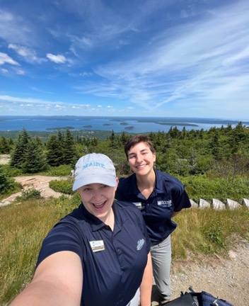 The author and another participant take a selfie while walking on a trail. Grass, trees, and the ocean is in the background. The sky is partly cloudy.